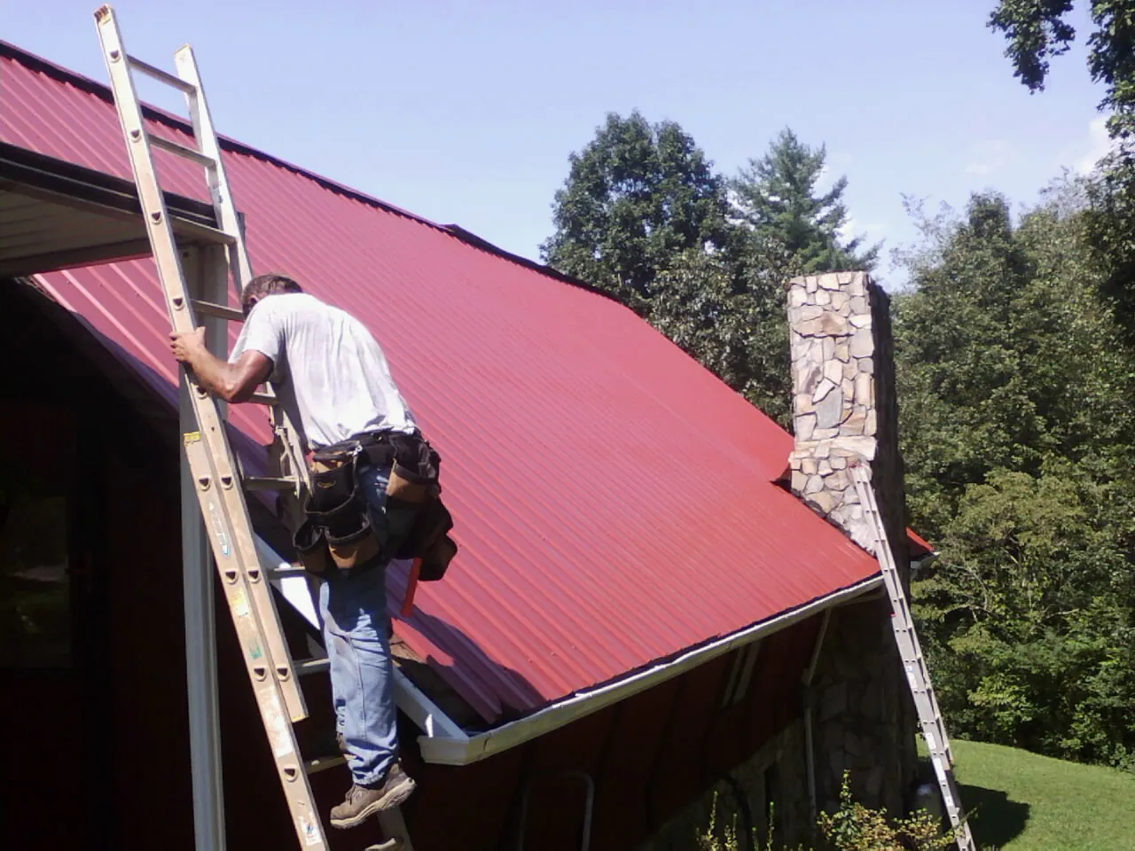 Roofer on a ladder beside a red metal roof and stone chimney — Soffit & Fascia Repair by Roofing Brothers in Kansasville